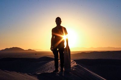 Silhouette man standing with sandboard in desert during sunset