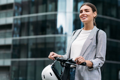 Smiling woman holding a handlebar of electric push scooter while standing at glass building	