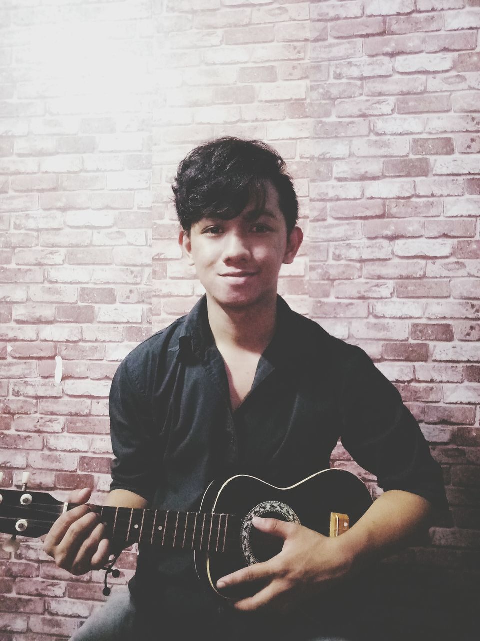 one person, brick, brick wall, wall, young adult, young men, wall - building feature, real people, looking at camera, portrait, front view, music, lifestyles, casual clothing, musical instrument, guitar, arts culture and entertainment, string instrument, leisure activity, musical equipment, musician, plucking an instrument