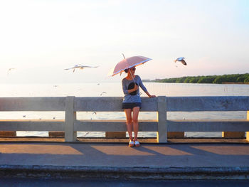 Woman holding umbrella while leaning on railing at bridge over sea during sunny day