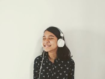 Smiling young woman standing against wall and listening to the music from headphone