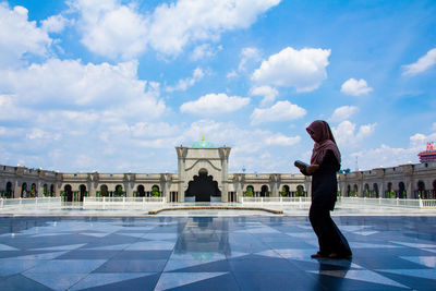 Full length of young woman holding holy book while walking at mosque against cloudy sky