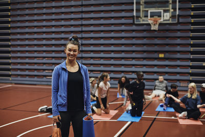 Portrait of smiling female coach standing with exercise mat against students in basketball court