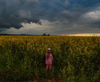 Rear view of girl standing at rape field against storm clouds