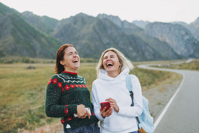 Happy young women travellers using mobile on road against beautiful mountain landscape