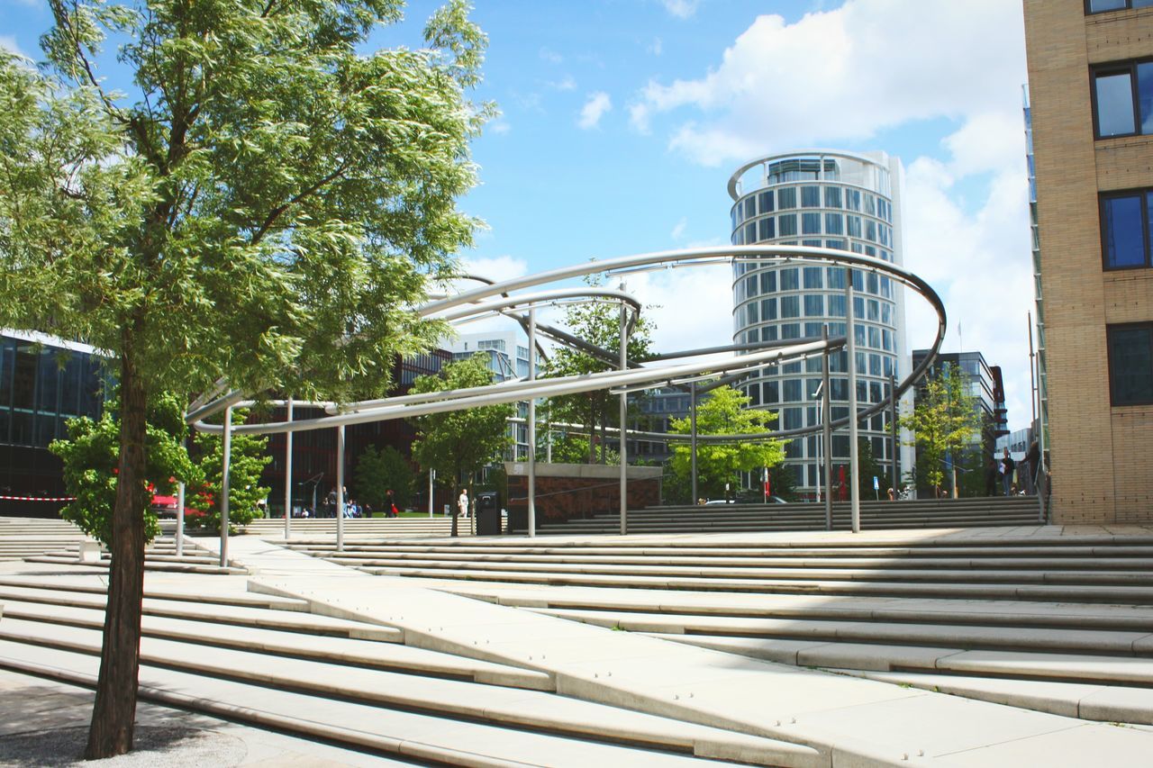 architecture, built structure, building exterior, tree, sky, glass - material, city, low angle view, building, modern, railing, day, window, outdoors, sunlight, metal, growth, office building, no people, cloud