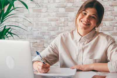 Smiling teenage student writing on book at home