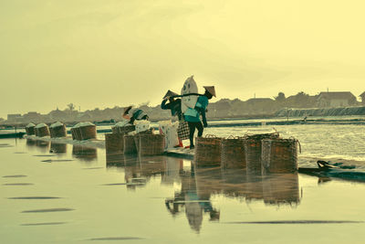 People working by lake against sky during sunset