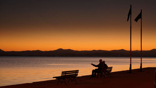 Silhouette man sitting by lake against orange sky