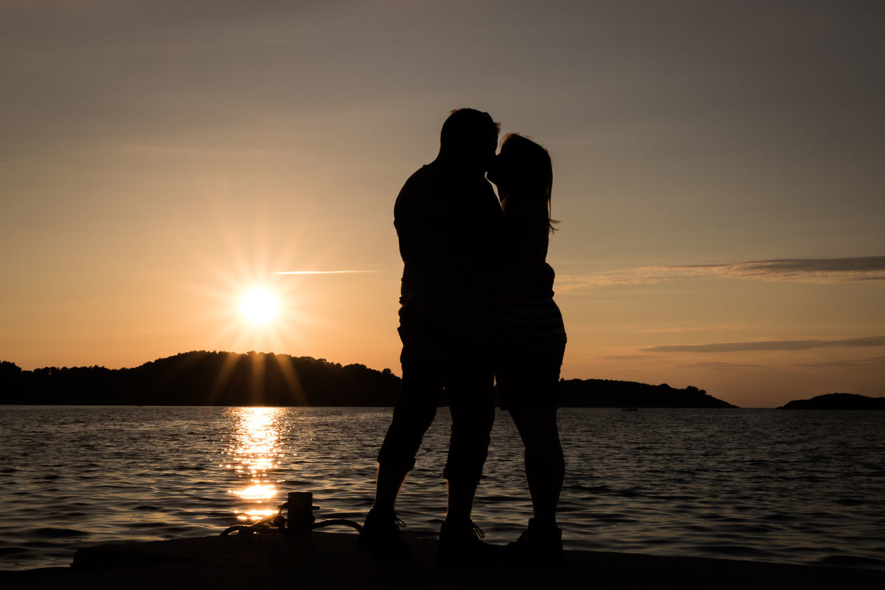 SILHOUETTE FRIENDS STANDING AT SEA SHORE AGAINST SKY DURING SUNSET