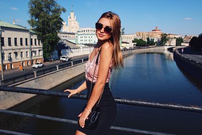 Portrait of young woman with sunglasses standing on bridge over river in city