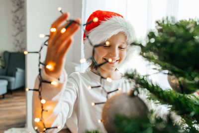 Charming girl in a santa claus hat decorates the christmas tree at home with a garland with lights. 