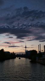 Silhouette buildings by river against sky at sunset