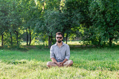 Young handsome european man in casual clothing and sunglasses sitting on a grass in summer park.