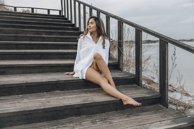 Woman sits barefoot on wooden staircase near lake, relaxed, white shirt dress, travel