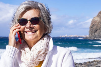 Smiling senior woman talking on mobile phone while standing against sea