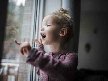 Girl with mouth open looking through window while standing at home