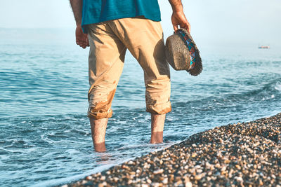 Male legs walking on pebble beach along the shore near the water with waves, low section.