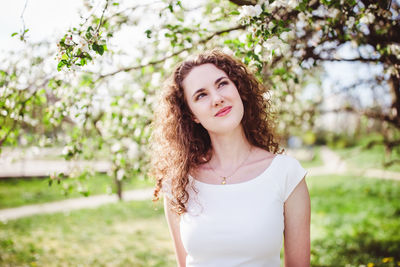 Happy curly hair young woman in a white t-shirt under the blooming apple tree. smiling, thinking