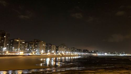Illuminated buildings by sea against sky at night