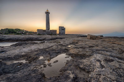 Lighthouse by sea and buildings against sky during sunset