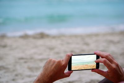 Cropped image of man photographing sea at beach on mobile phone