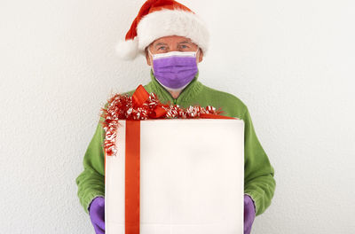 Portrait of smiling senior man wearing mask holding christmas box standing against wall