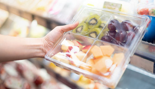 Female hand holding mix of fresh fruit in the transparent plastic box with fork in the supermarket.