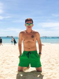 Portrait of shirtless man wearing sunglasses and shorts while kneeling at beach against sky during summer