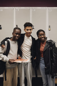 Portrait of smiling teenage boys standing with female friend against locker in school