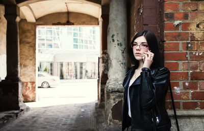 Sad young girl in black jacket and jeans stands in old arch on city street on summer day