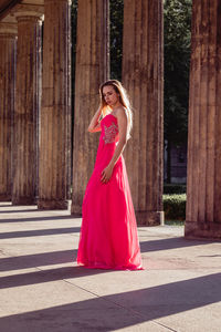 Full length side view of young woman in pink evening gown standing at colonnade