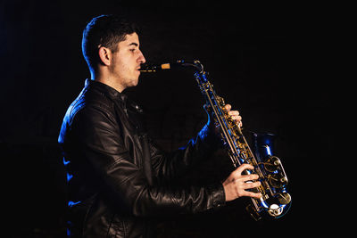 Side view of young man playing saxophone against black background