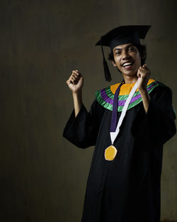 Portrait of smiling man wearing graduation gown standing against wall