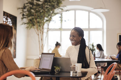 Happy female entrepreneur talking with colleague working on laptop at office