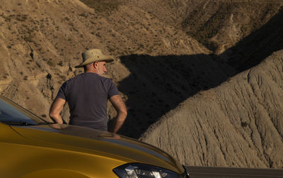 Adult man in cowboy hat standing against car on desert against mountain. almeria, spain