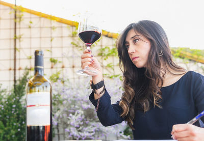 Hispanic woman inspecting red wine in glass and writing down data during degustation session on restaurant terrace on summer day