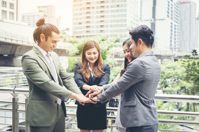 Smiling colleagues stacking hands while standing against buildings in city