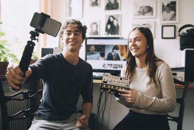 Teenage boy taking selfie on mobile phone in monopod with friend holding sound mixer at home