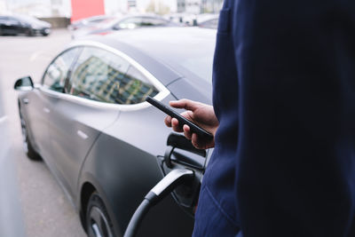 Businessman using smart phone standing by car at electric vehicle charging station