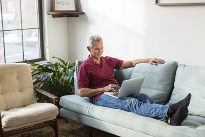 Full length of mature man using laptop computer while resting on sofa in living room at home