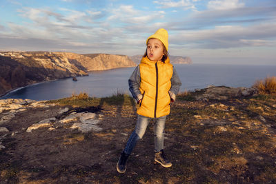 Boy child in a yellow hat and vest stands on top of a mountain by the sea in autumn