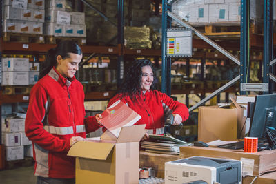 Pleased female employees in red uniform searching packages and collecting parcels in big modern warehouse