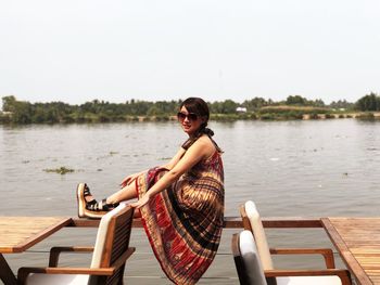 Side view of young woman sitting on railing by lake against clear sky