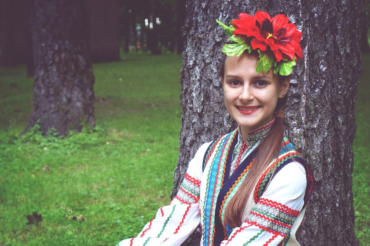 looking at camera, portrait, front view, lifestyles, person, standing, leisure activity, casual clothing, young adult, smiling, young women, red, focus on foreground, flower, tree, park - man made space, grass