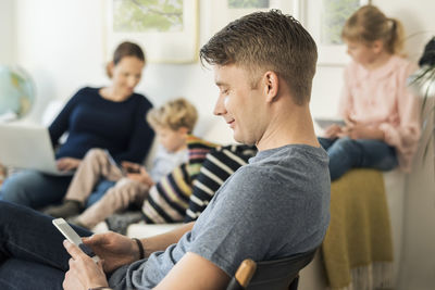 Side view of man using phone with family sitting on sofa at home