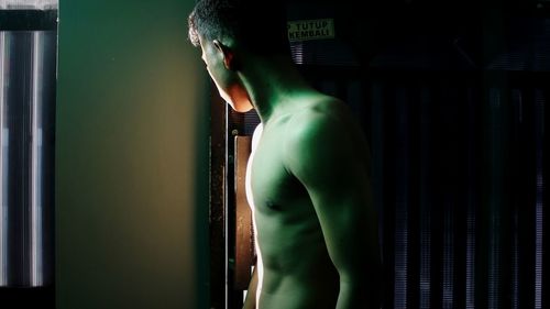 Side view of shirtless man standing by window