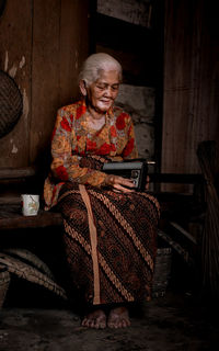 Full length of senior woman holding vintage radio while sitting outdoors