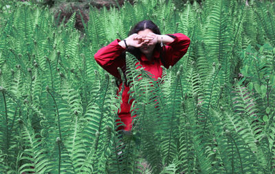 Young woman covering her eyes while standing amidst ferns on field