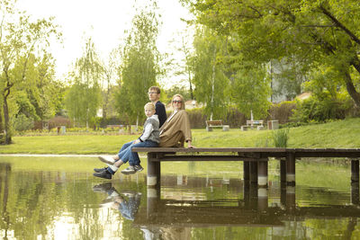 Caucasian family mother, father and son sit on wooden dock enjoying lake view in summer time. family